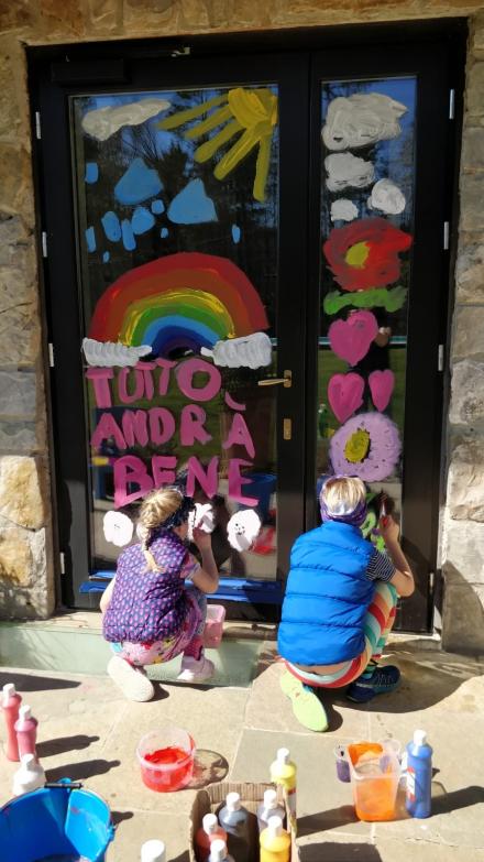 2 persons painting "Tutto andra bene", flowers, hearts and rainbow on a door. In the foreground painting glasses and buckets. 