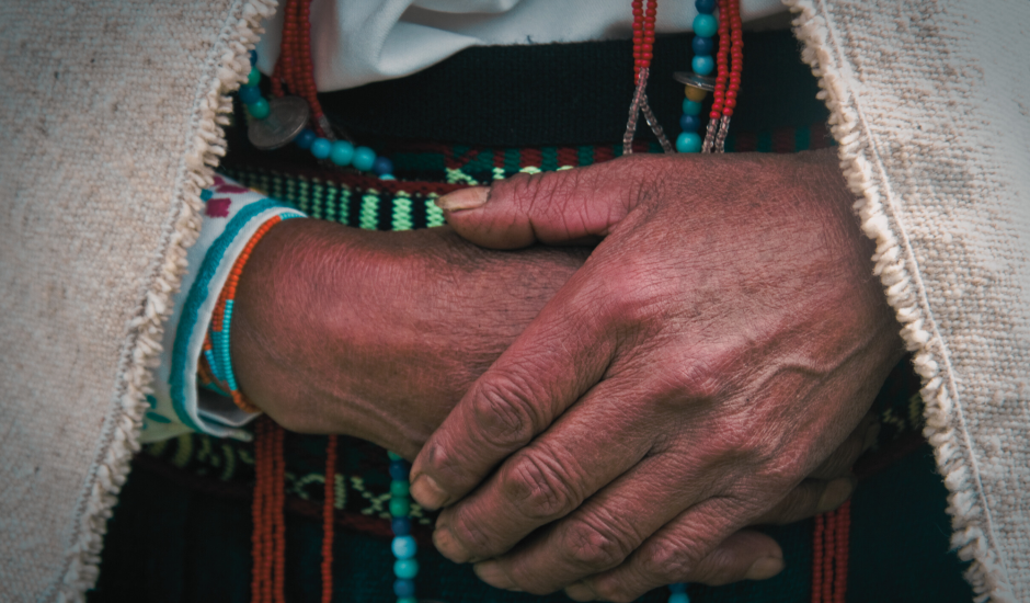 An indigenous person in colorful clothing and accessories, the camera is zoomed on the hands that are crossed. 