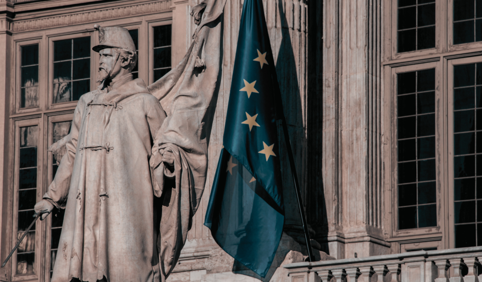 A classical building, in front of it, there is a sculpture of a man in official clothing. There is also an EU flag in front of the building. 