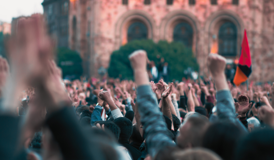 Many hands lifted on the photo, with some unrecognizable flag in the background and also a brown brick building. It portrays a protest. 
