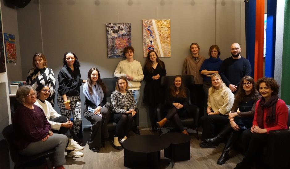 A group of people (15) sitting in chairs, some of them standing. 3 artworks (abstract) in the background)