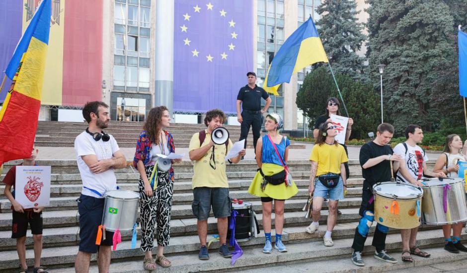 A group of demonstrators stands on the steps of a government building during a public rally. One person speaks into a megaphone while others hold papers or play drums. Large Ukrainian, European Union, and Moldovan flags hang behind them, with additional Ukrainian and Moldovan flags being held by participants. A security guard stands on the steps behind the group, and trees frame the building in the background.