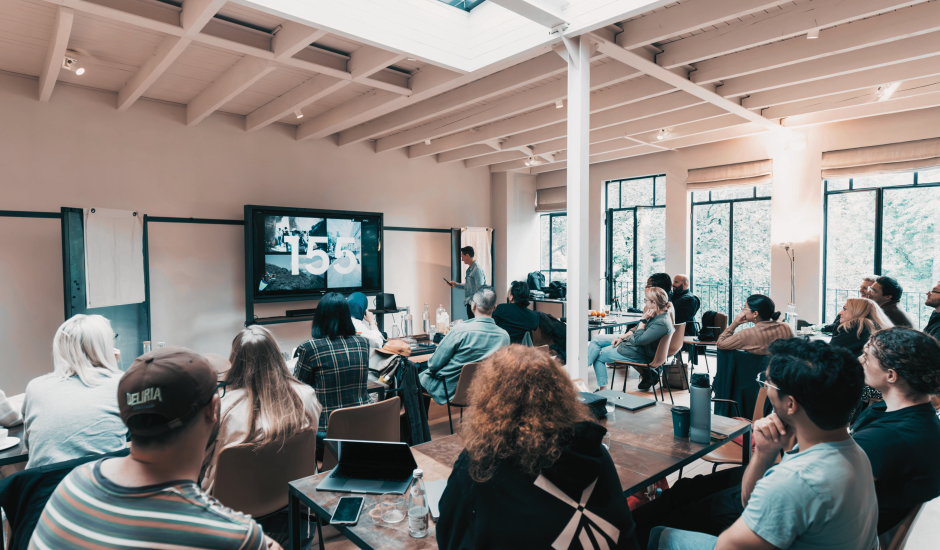 A group of people in a smaller meeting room, there is a speaker in the front and they are all looking at a screen