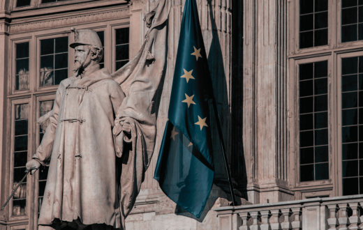 A classical building, in front of it, there is a sculpture of a man in official clothing. There is also an EU flag in front of the building. 