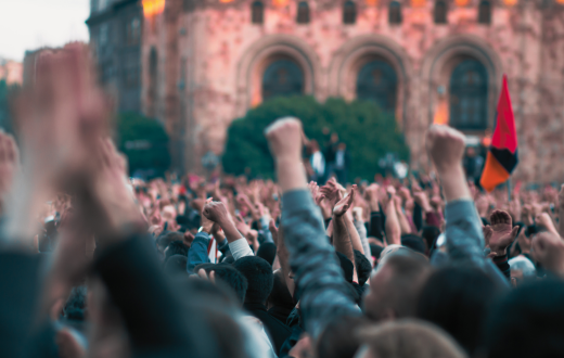 Many hands lifted on the photo, with some unrecognizable flag in the background and also a brown brick building. It portrays a protest. 