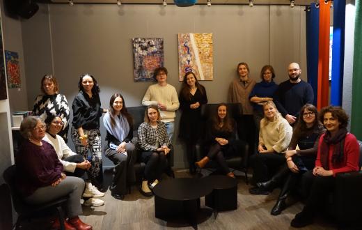 A group of people (15) sitting in chairs, some of them standing. 3 artworks (abstract) in the background)