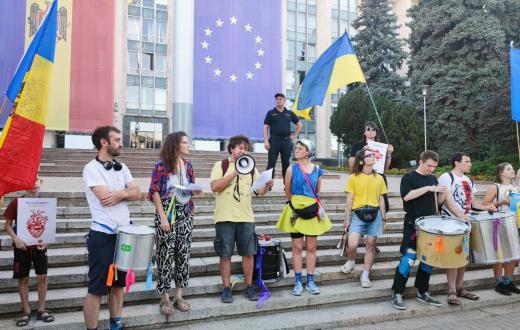 A group of demonstrators stands on the steps of a government building during a public rally. One person speaks into a megaphone while others hold papers or play drums. Large Ukrainian, European Union, and Moldovan flags hang behind them, with additional Ukrainian and Moldovan flags being held by participants. A security guard stands on the steps behind the group, and trees frame the building in the background.
