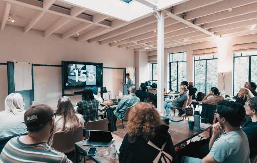 A group of people in a smaller meeting room, there is a speaker in the front and they are all looking at a screen